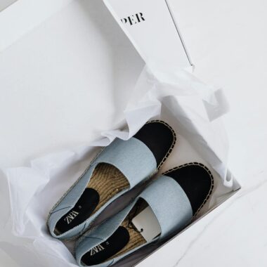 Top view of fashionable espadrilles in an open white cardboard box on a marble surface.